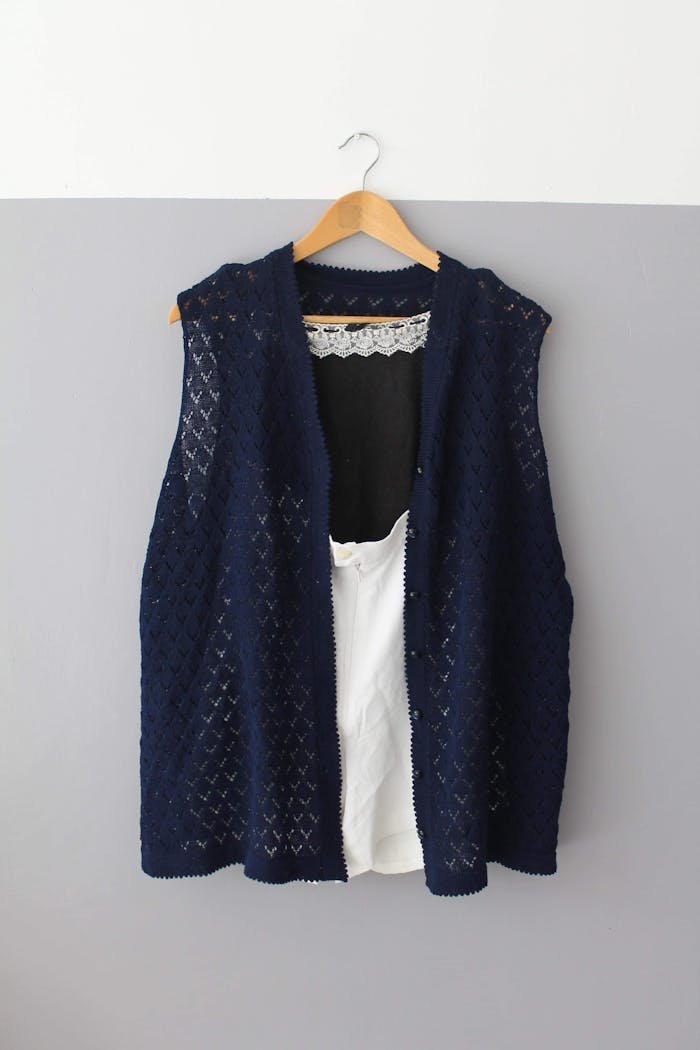 A dark blue crochet vest displayed on a wooden hanger against a neutral wall.